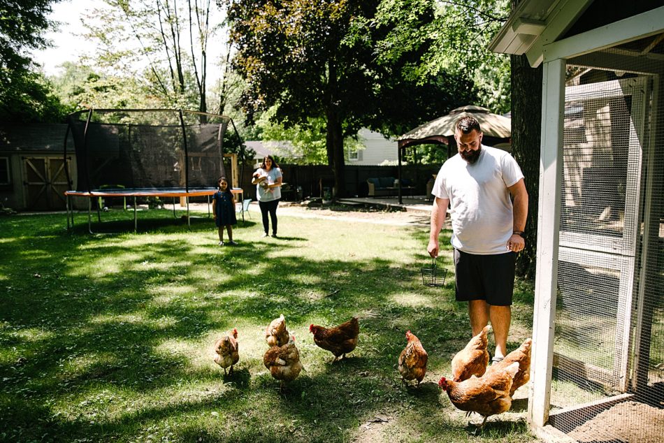Mom and children stand in background while father handles chickens in backyard coop
