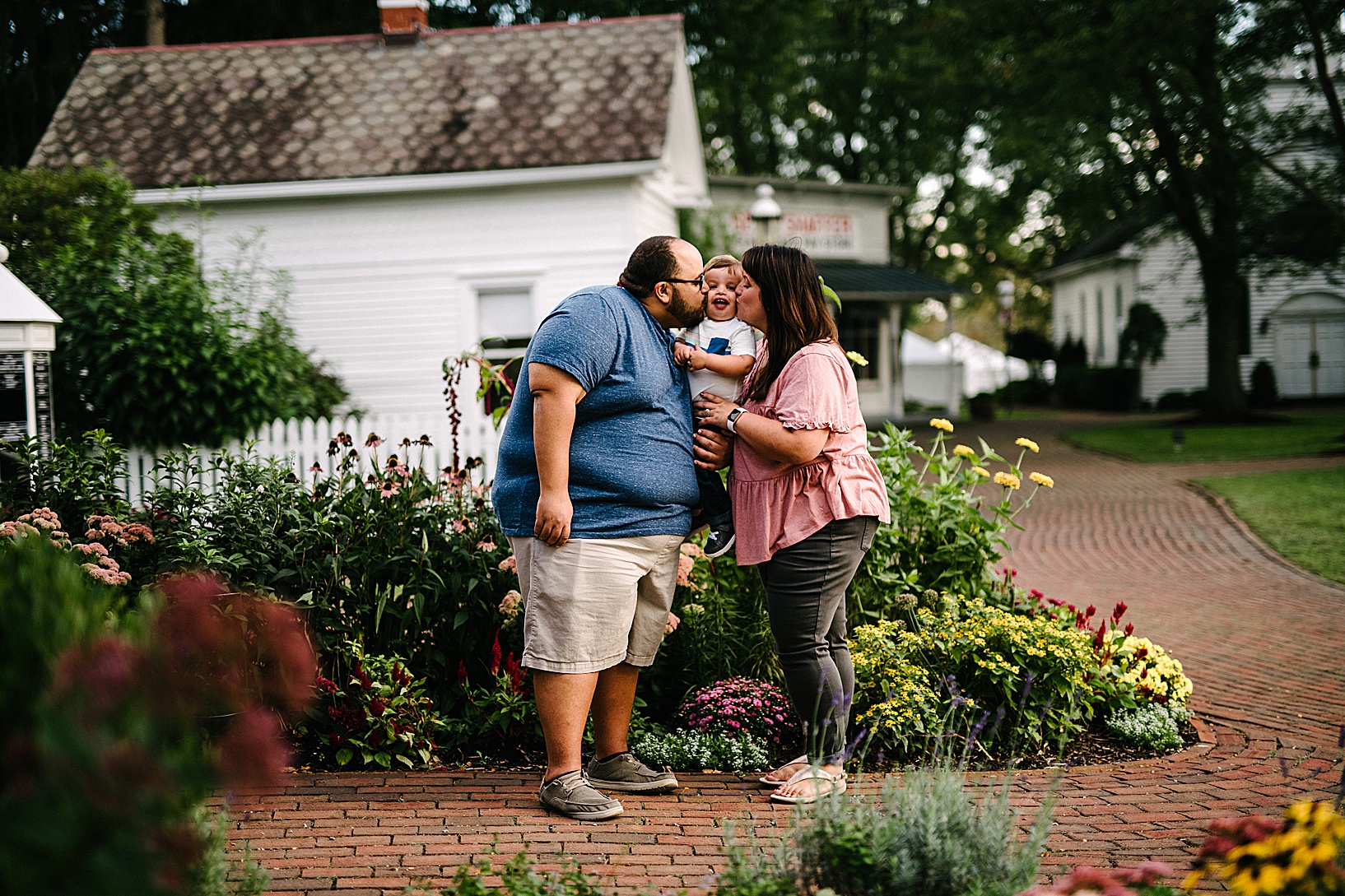 Man and woman hold their son between them while kissing him on the cheek with old house and wildflowers in the background at Western Village Reserve