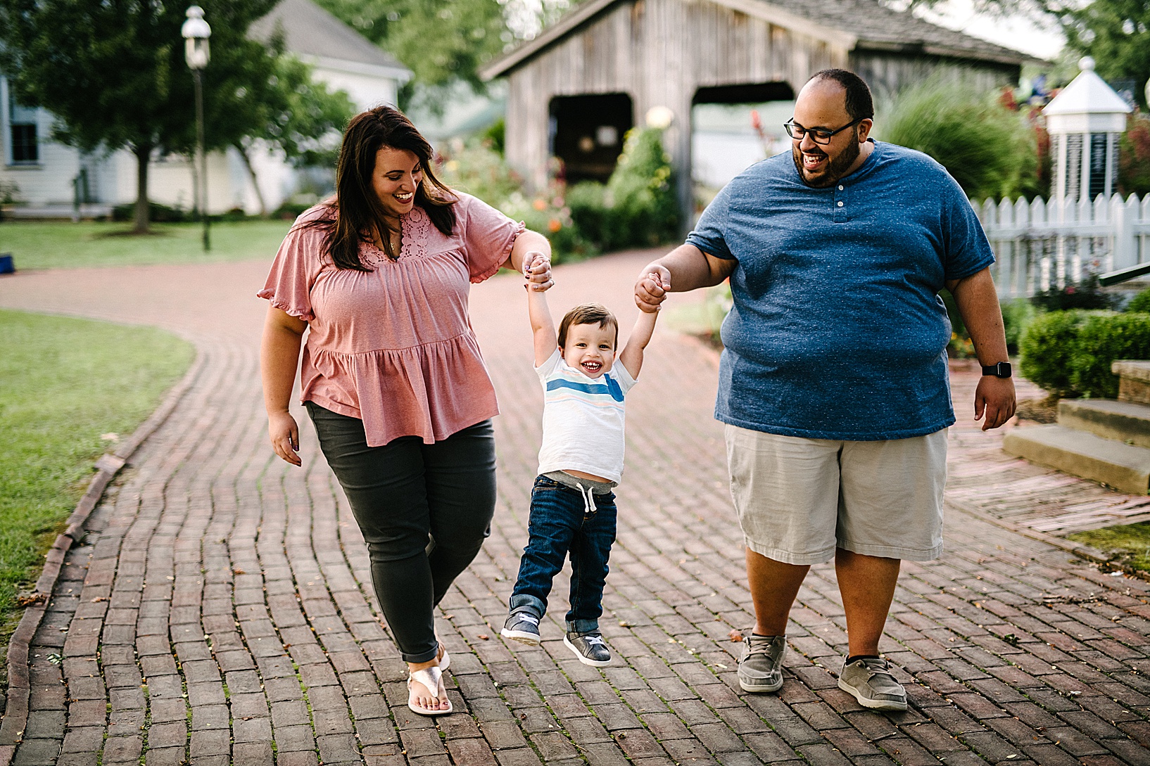 Woman in pink shirt and man in blue shirt walk along a brick pathway and swing their young son by the hands between them while they smile