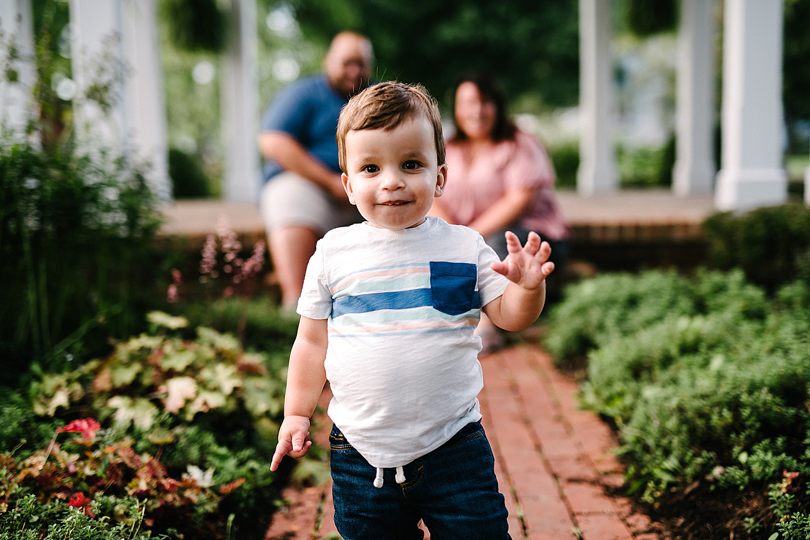 Young boy walks down a brick pathway smiling while his parents watch him in the background.