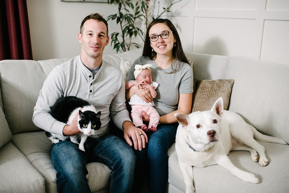 Woman and husband pose on couch with their newborn baby, white dog, and black and white cat.