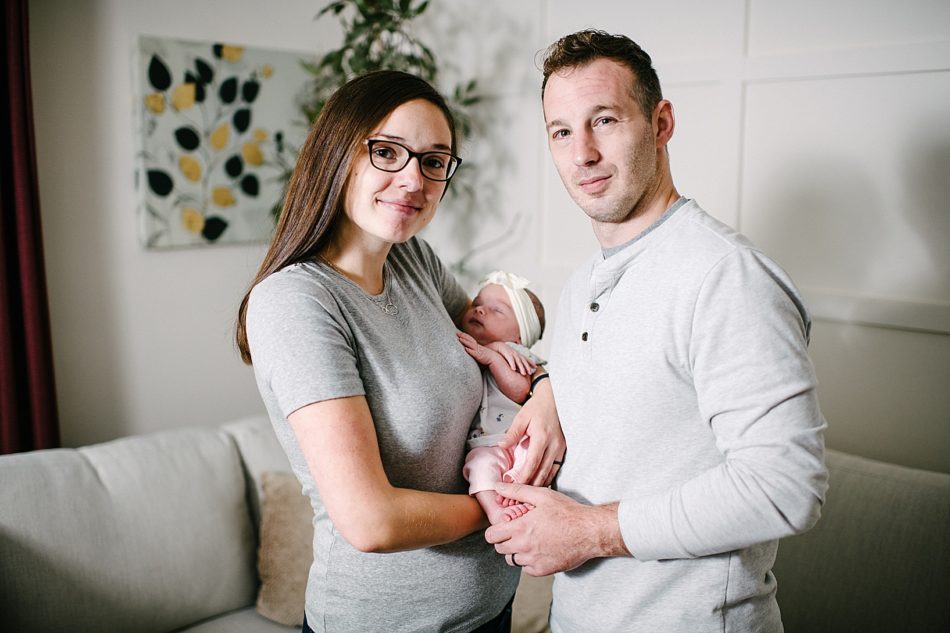 Woman and man smile at the camera while holding newborn baby wearing white linen bow