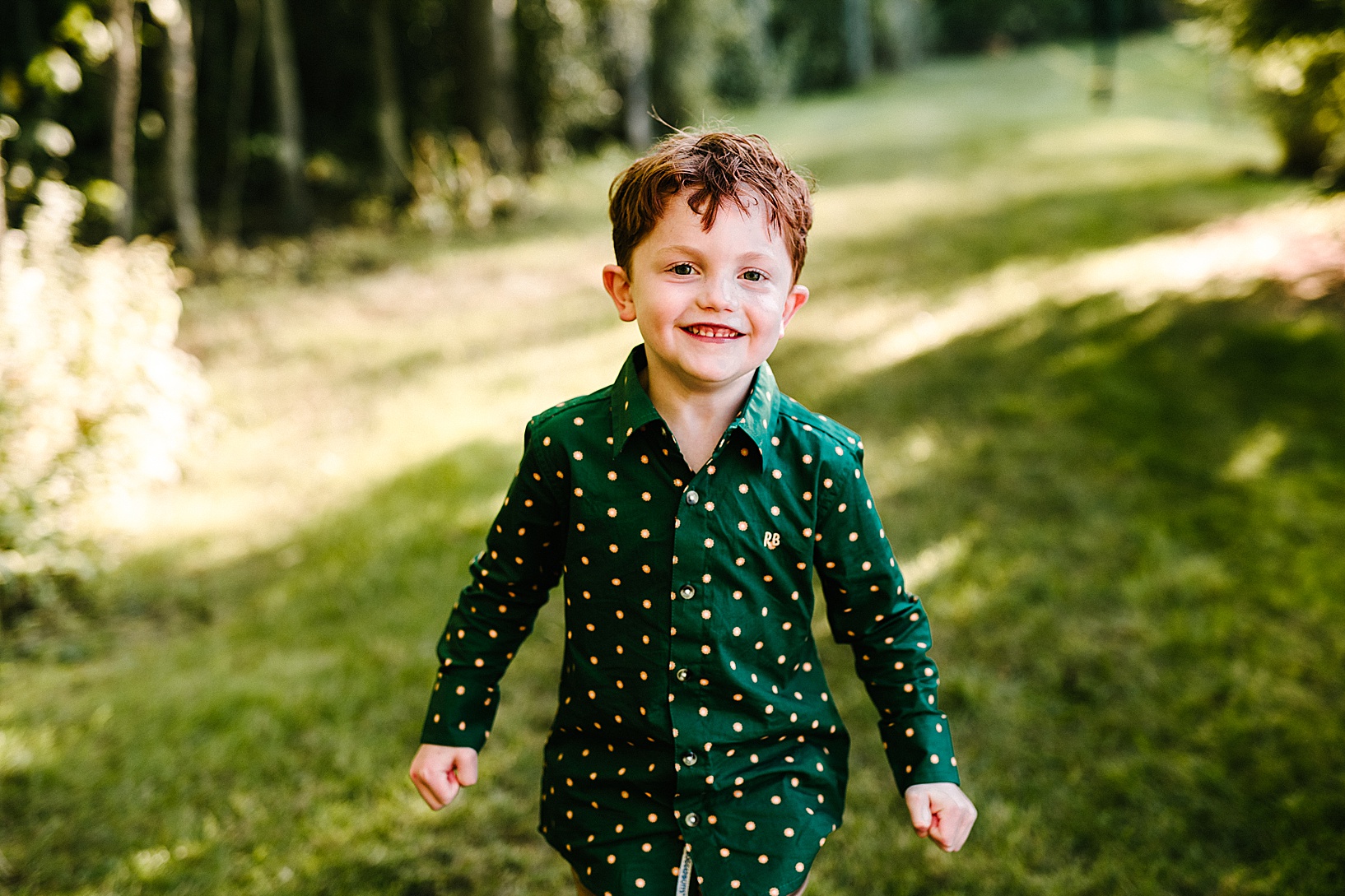 Young Red headed boy in a green polka dot shirt smiles at the camera with a grass field in the background during fall family session