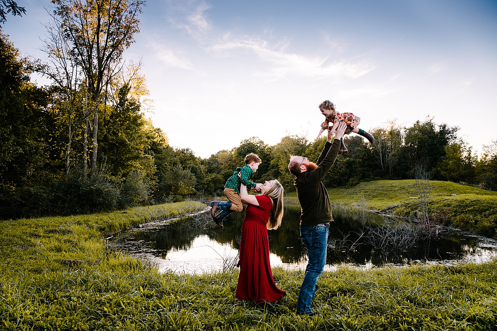 Blonde woman and husband stand back to back in front a dreamy lake while lifting their son and daughter high into the air during fall family session
