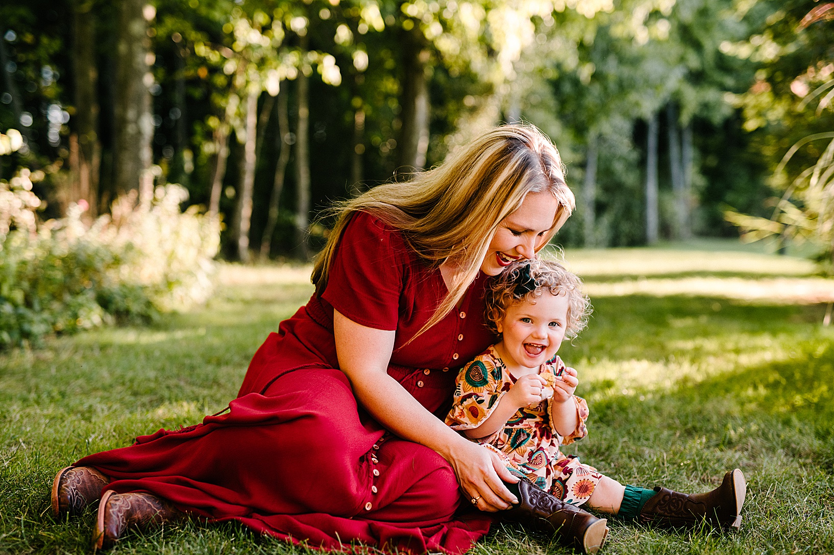 Blonde woman in long red dress sits on the grass cuddling with her young daughter during fall family session