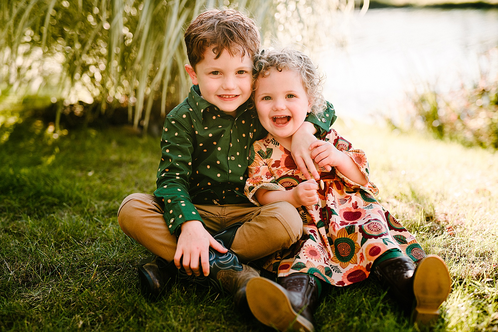 Young boy puts his arm around his toddler sister sitting on the grass while they both wear cowboy boots