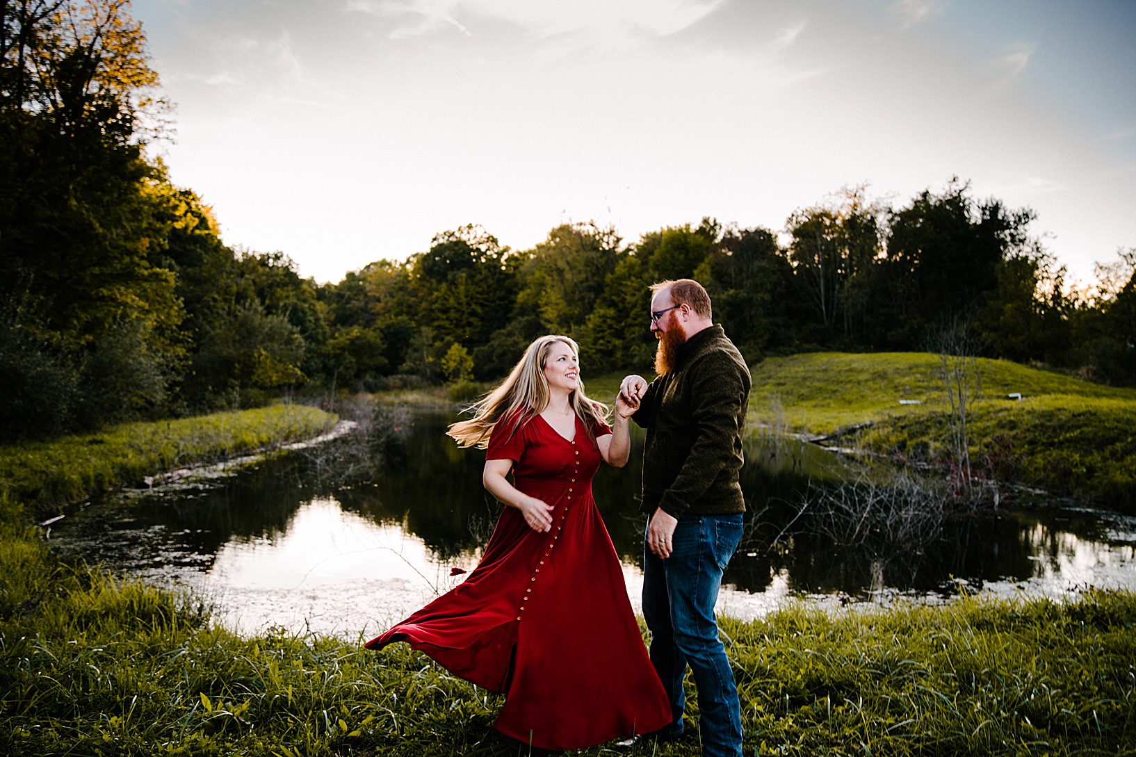Husband twirls blonde wife in red dress in front of dreamy lake fall family session