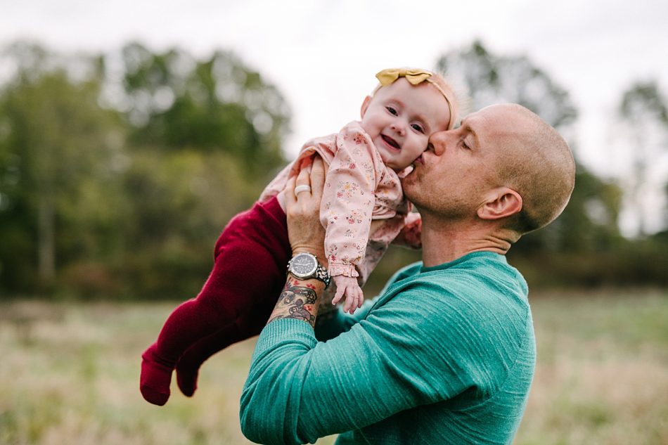 Bald man in teal shirt holds his baby daughter in the air and kisses her on the cheek as she holds a huge smile.