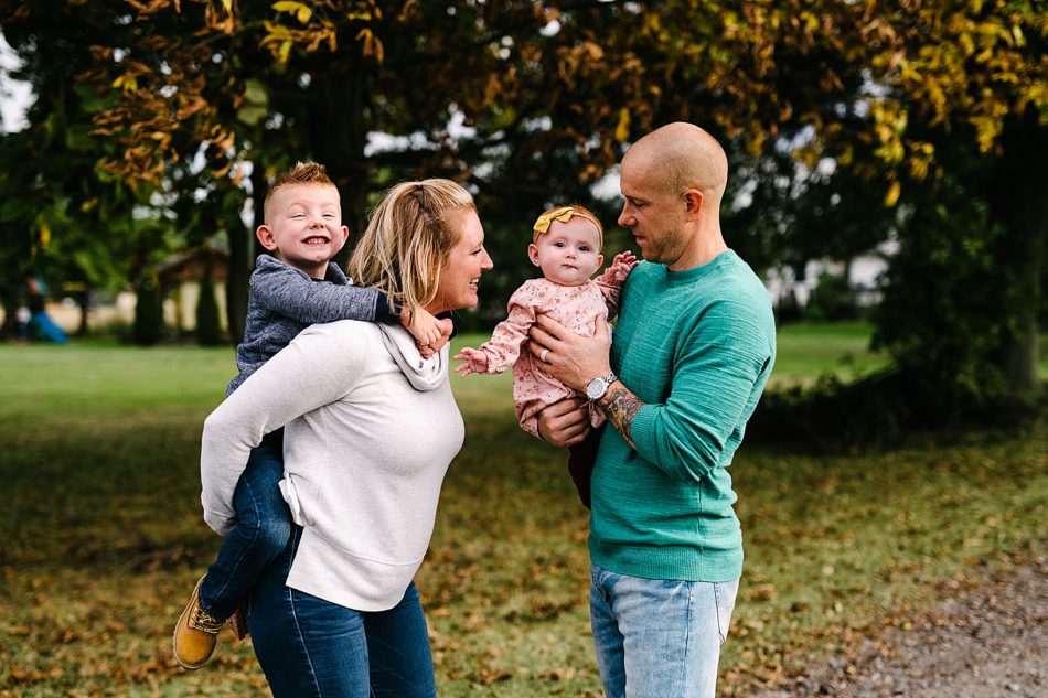 Blonde woman in white sweater carries her young son on her back while man in teal shirt carries their baby daughter in front of gorgeous fall foliage in North Jackson.