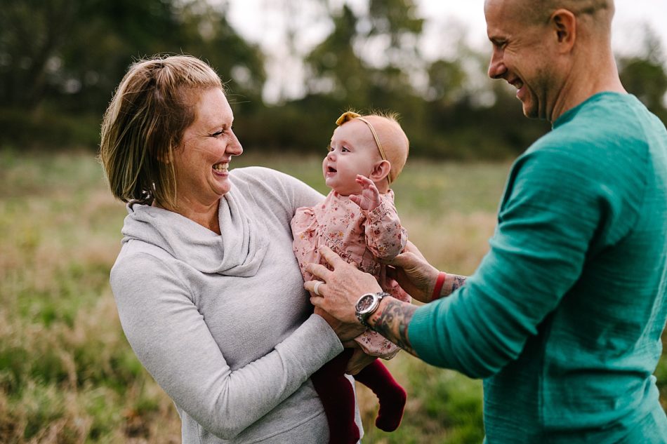 Mom smiles while holding her baby girl while dad holds her from behind.