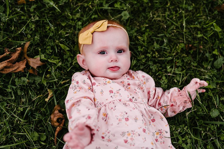 Baby girl in a pink floral dress with a big yellow head bow lays in the grass with her tongue sticking out.