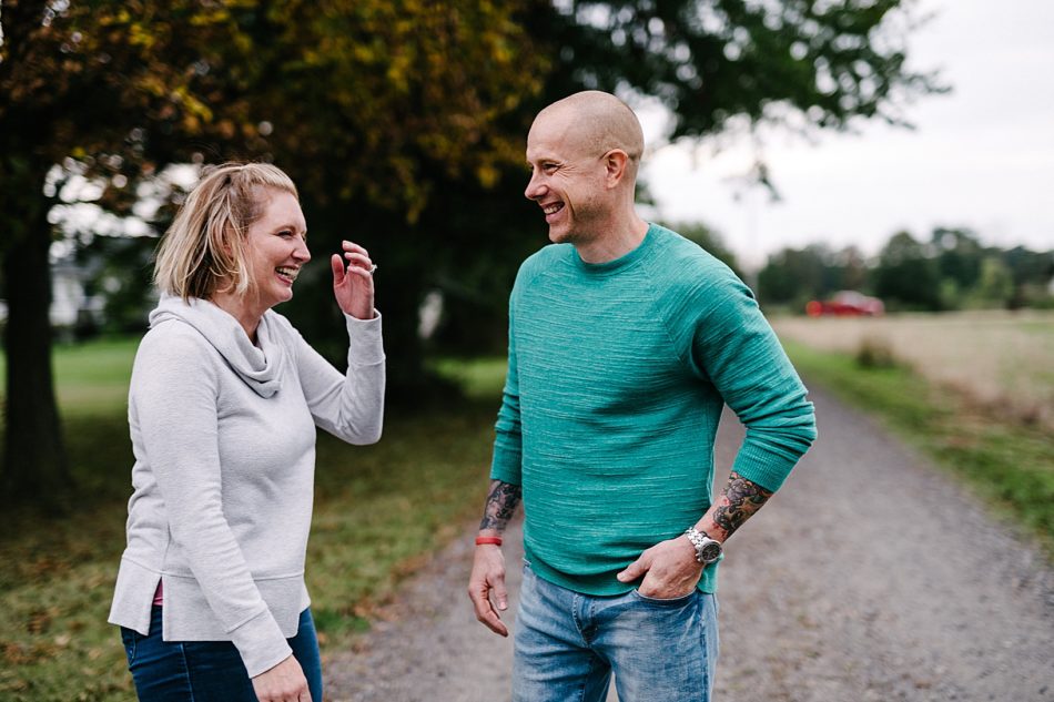 Blonde woman laughs and brushes her hair back while walking towards her husband in a teal shirt on standing on a gravel road