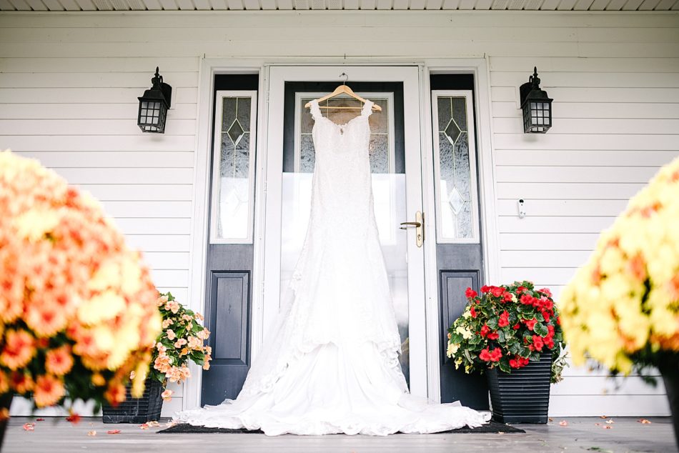 The wedding gown hanging from the screen door of a white house with a black door, black lights on each side of the door, and red and orange flowers on each side of the door