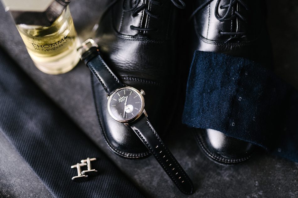 Groom accessories including black dress shoes with a navy polishing cloth, black and gold Shinola watch, gold cufflinks, Thomme cologne, and a black tie