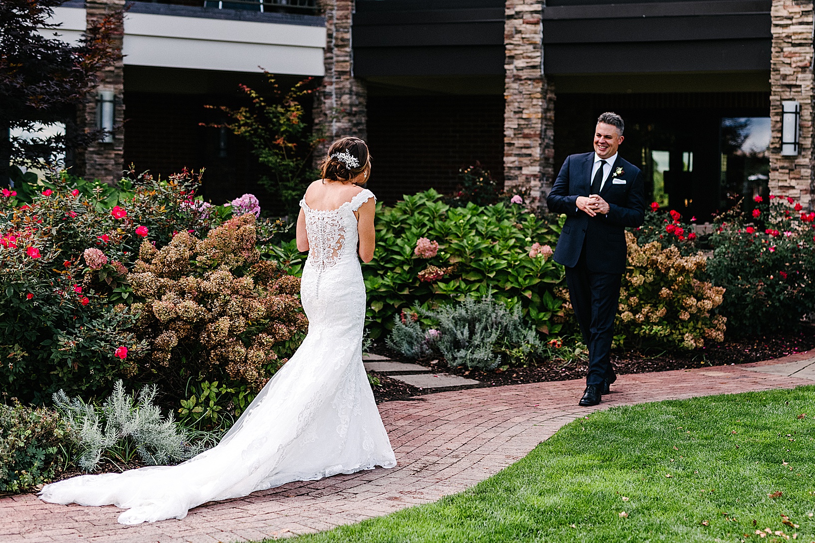 Groom smiles as he sees his bride for the first time, as she walks towards him down a red brick pathway