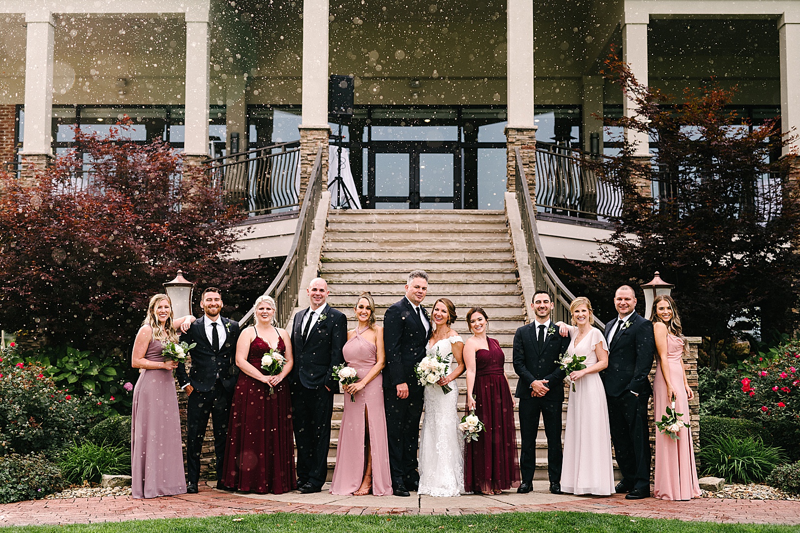 The wedding party all stands and smiles in front of the big steps of the Lake Club while light rain falls