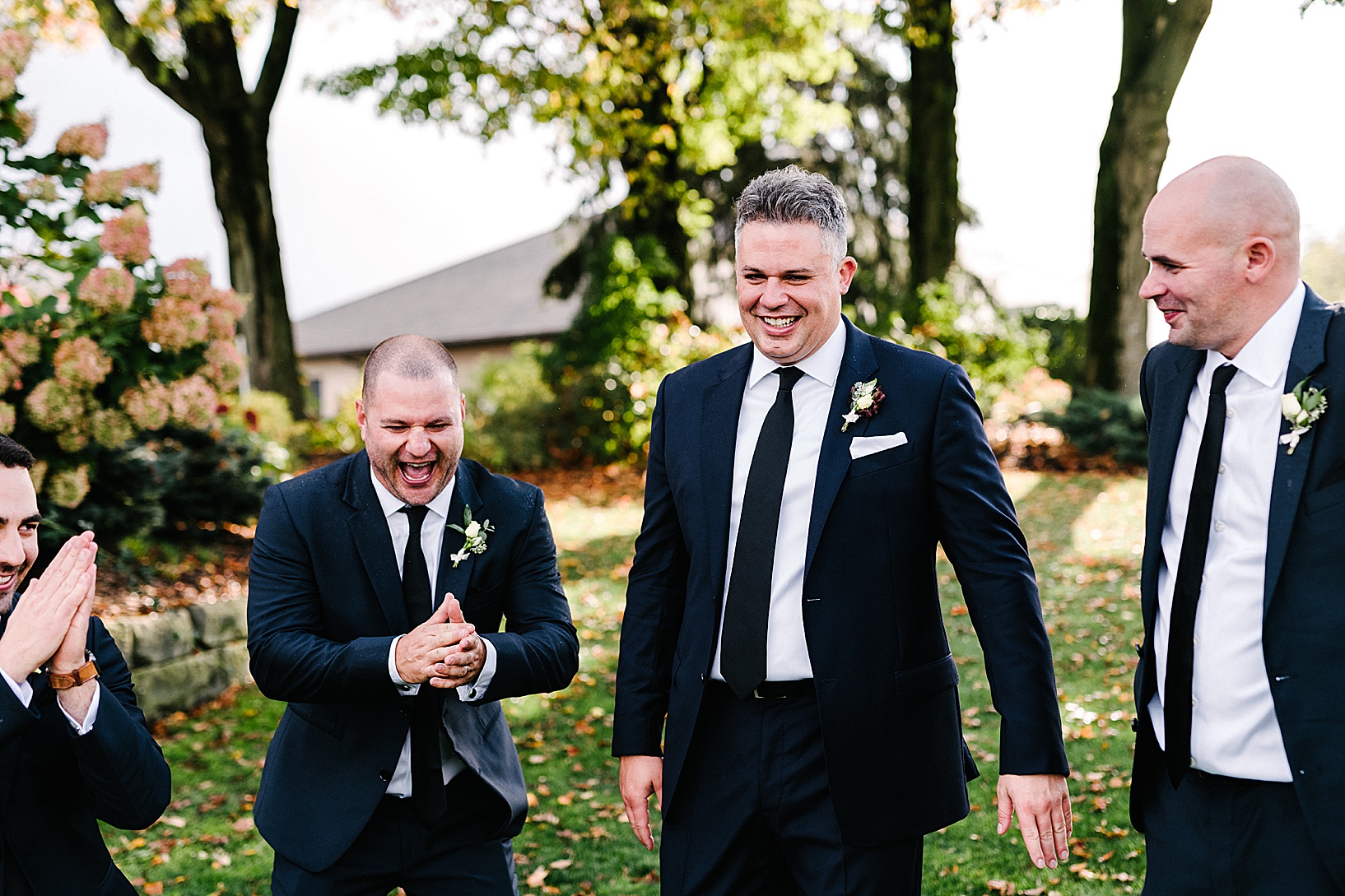 A groomsman claps his hands laughing while the groom smiles and laughs while standing on the lawn of the Lake Club.