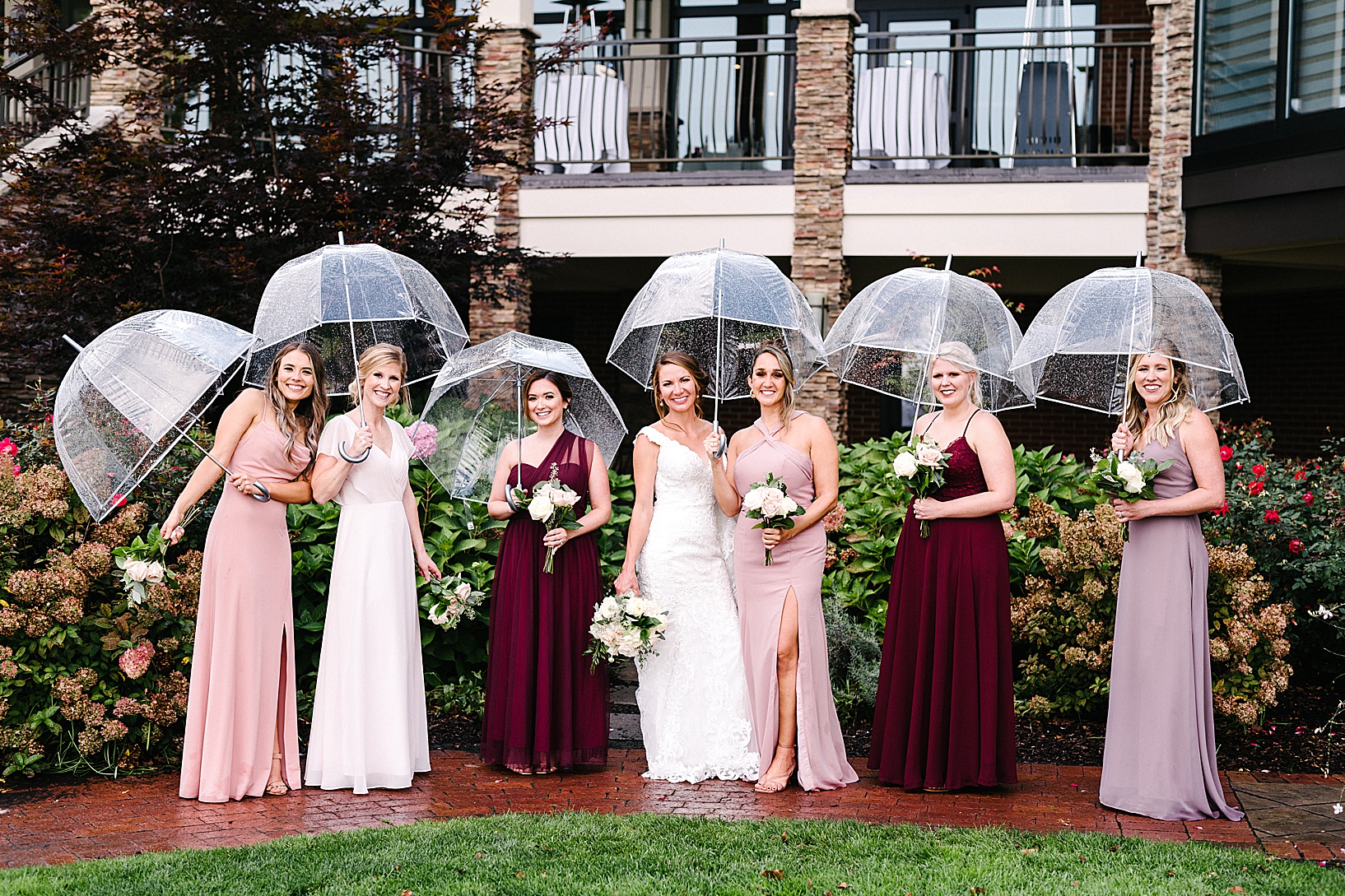 The bridal party and the bride standing on a brick pathway of the Lake Club, holding clear umbrellas.