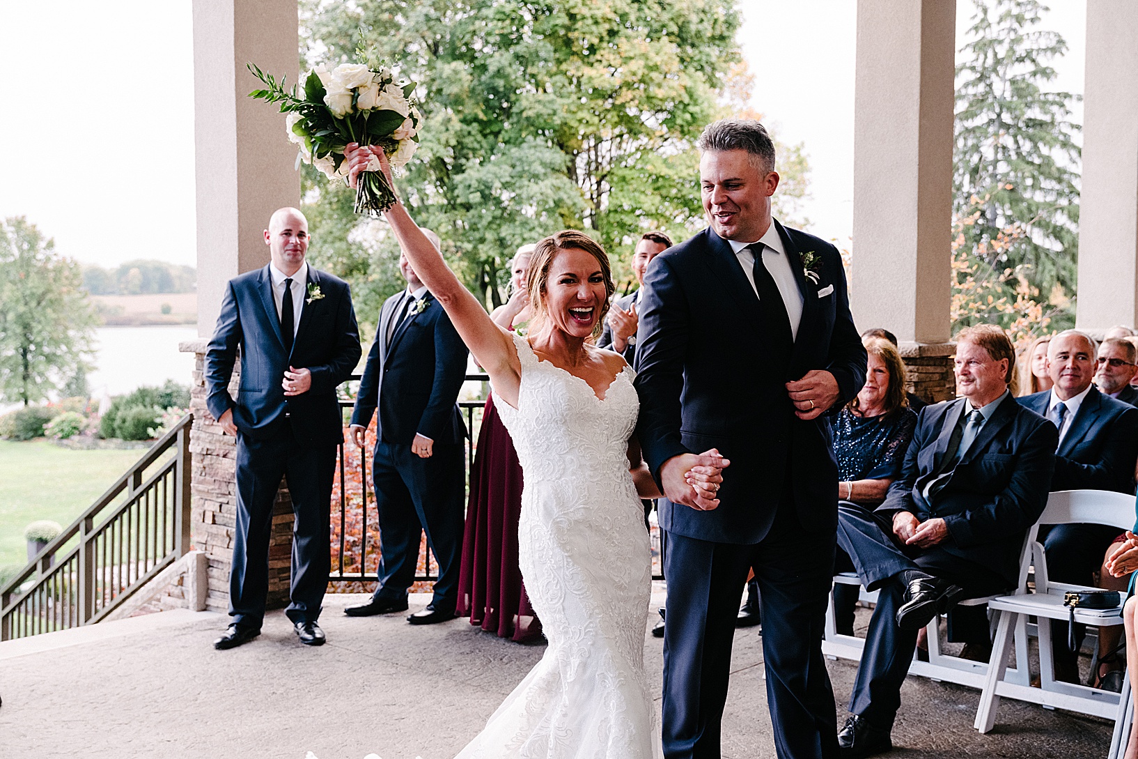 Bride holds her bouquet in the air during recessional while the groom holds her hand and smiles at her.