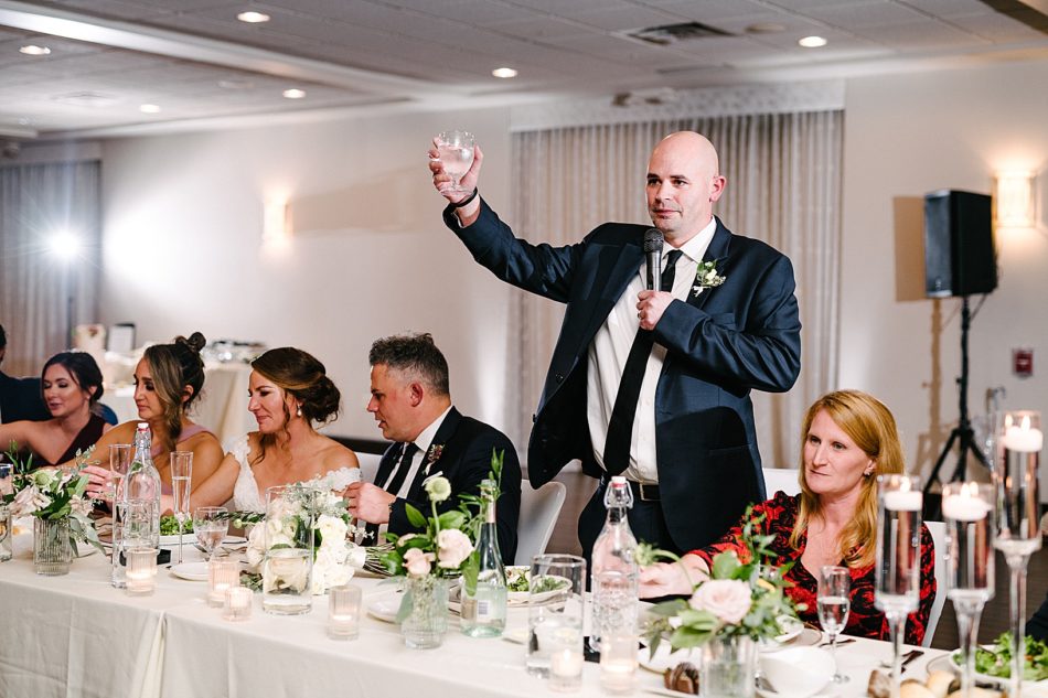 The best man raises a glass while holding a microphone during his speech.
