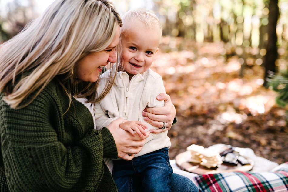 Blonde woman in chunky knit, dark green cardigan smiles and hugs her young blonde son as he smiles at the camera.