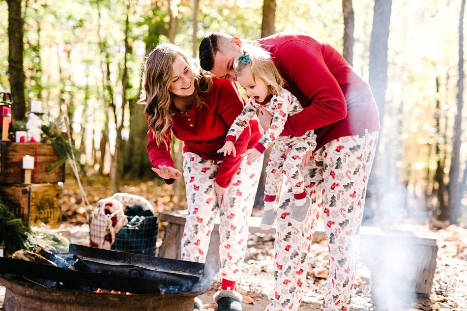 Family of three, woman, man, and a young blonde girl all wear matching christmas pajamas as dad holds daughter closer to the campfire to warm up her hands.