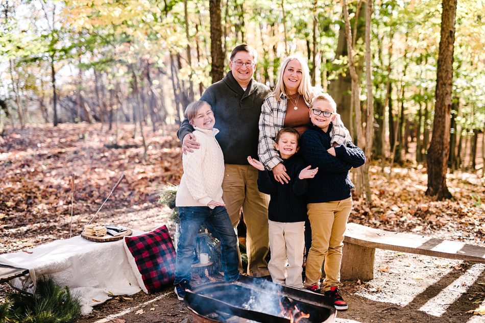 Family of five, a woman, man, and three young boys all stand behind a cozy campfire at Lamppost Farm with smores, pillows, and throws in the background.