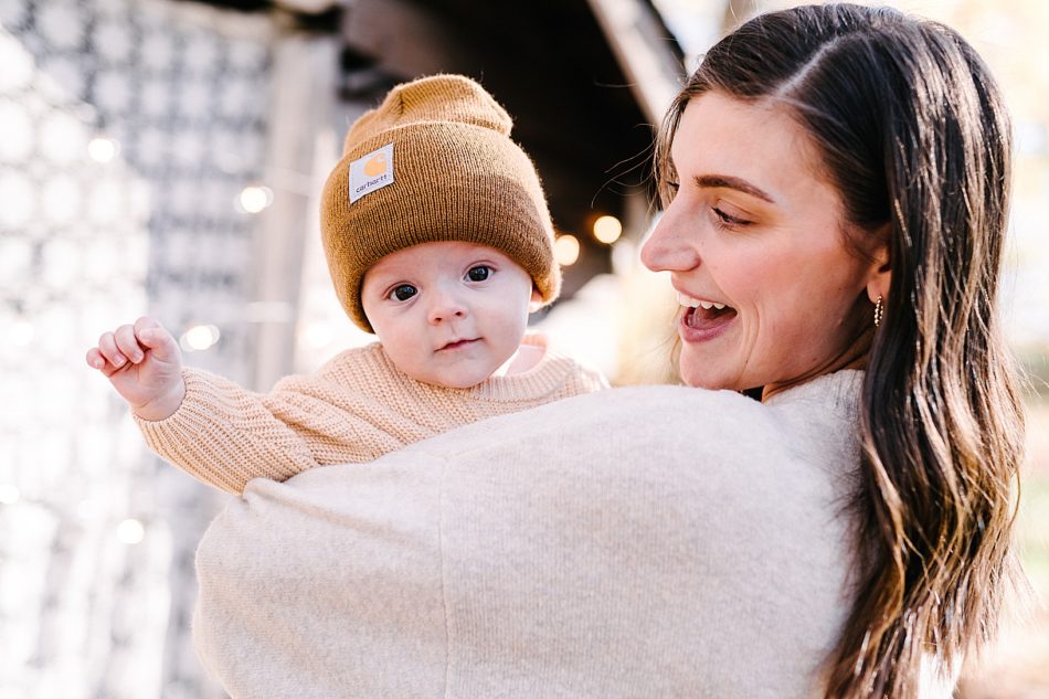 Brunette woman holds her baby son who is in a mustard colored hat and ivory knit sweater, and smiles at him as he looks over her shoulder at the camera.