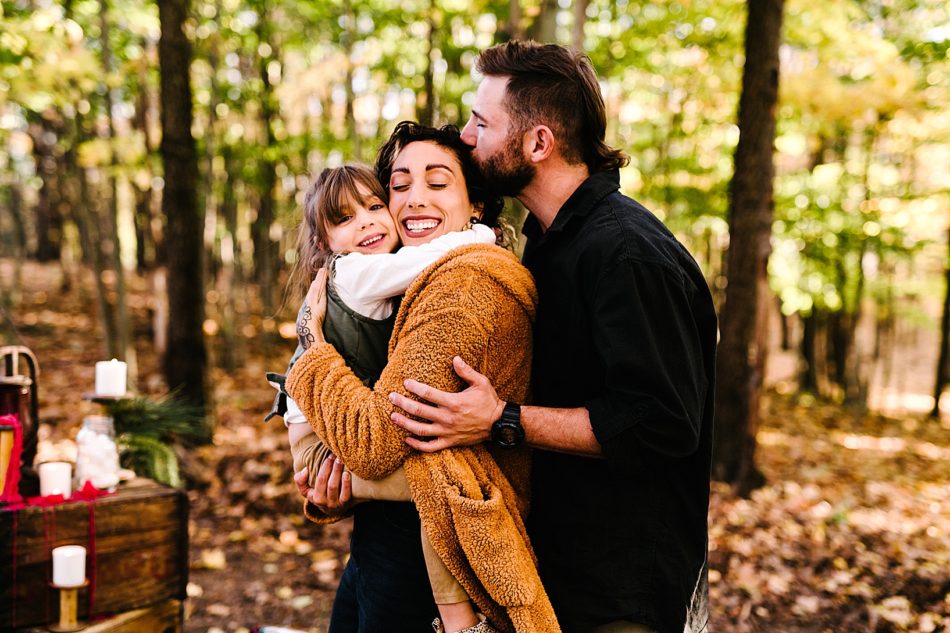 Brunette woman in fuzzy orange cardigan cuddles her daughter while her bearded husband kisses her head and hugs her from behind.