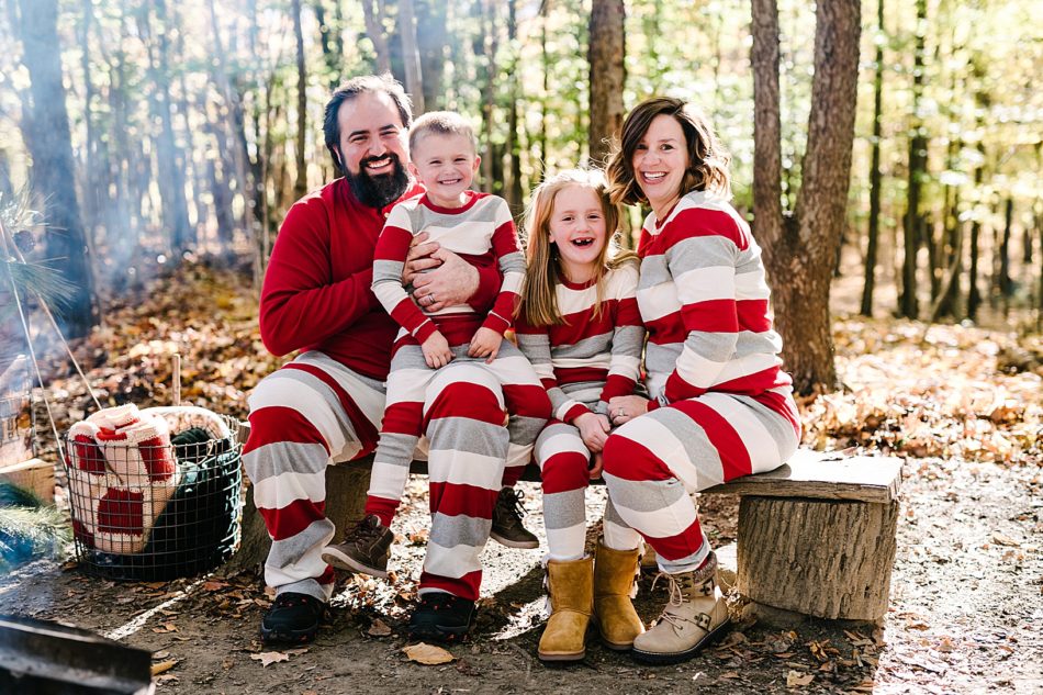 Family of four wearing matching gray, white, and red pajamas gather on tree stump bench with a basket of blankets and wooden foliage of Lamppost Farm in the background.