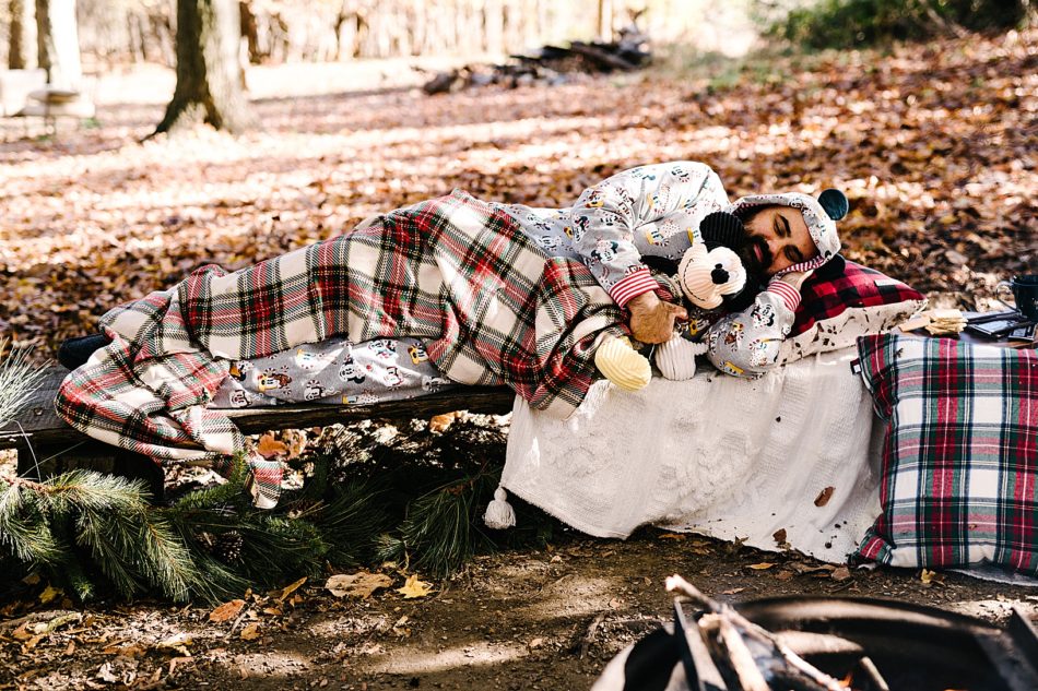 Bearded man in Mickey Mouse themed, hooded onesie sleeps on bench with flannel blanket and Mickey Mouse stuffed animal.