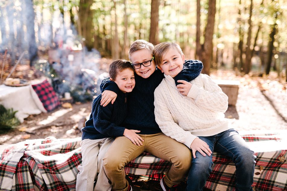 Three brothers sit on a flannel blanket covered bench with the middle one's arms wrapped around his brothers necks.