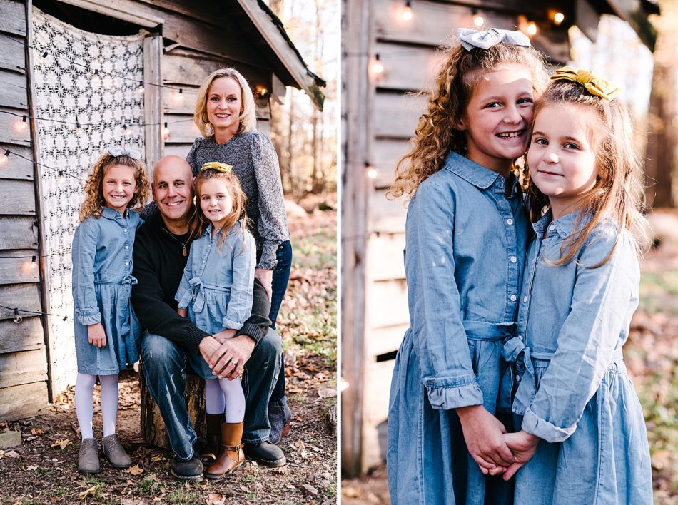 Two young blonde girls in matching denim dresses and hair bows stand cheek to cheek and smile in front of a wooden cabin at Lamppost Farm.