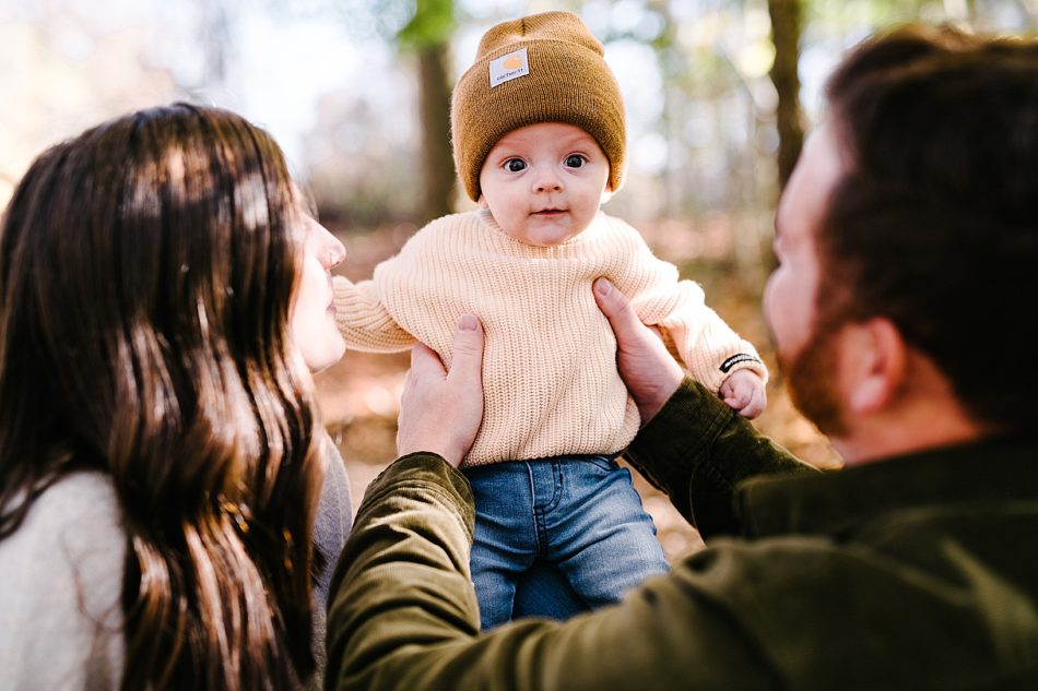 Man holds up baby wearing a mustard hat, an ivory knit sweater, and blue jeans while dad and mom smile at him.