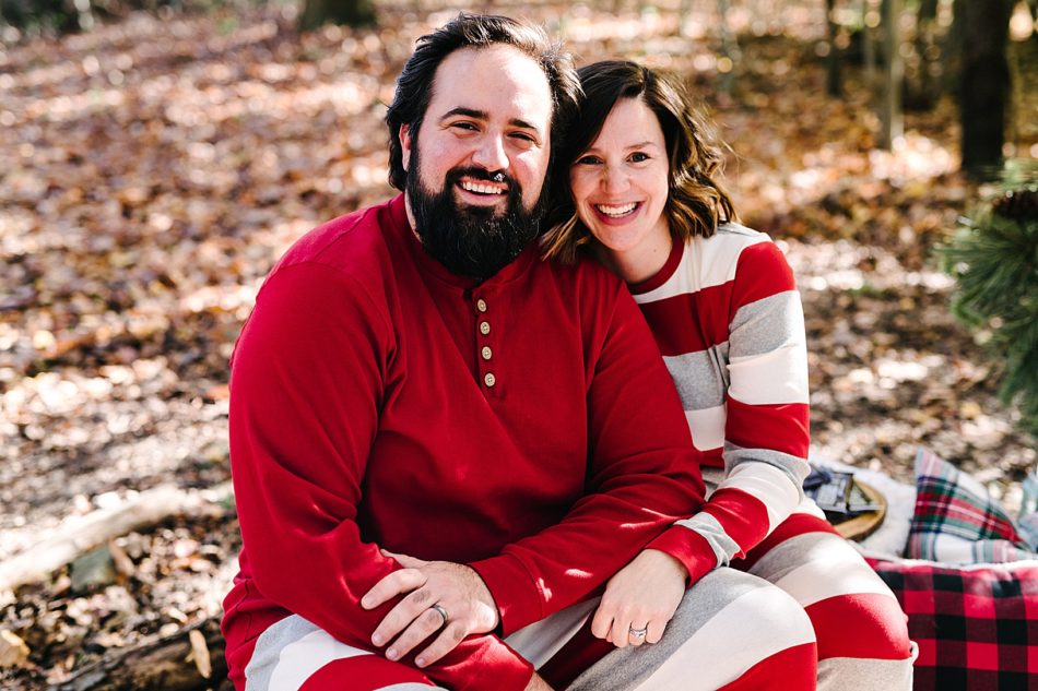 Bearded man and brunette woman sit close and smile as they wear matching gray, white, and red striped pajamas.
