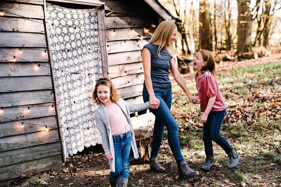 Woman laughs and holds hands of two young girls in front of the cozy wooden cabin at Lamppost Farm.