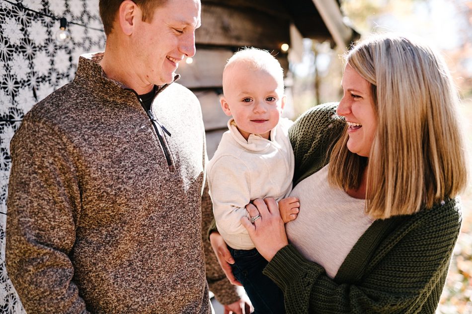 Man in speckled sweater and blonde woman in green knit cardigan smile and both look at their blonde son as he smiles at the camera.