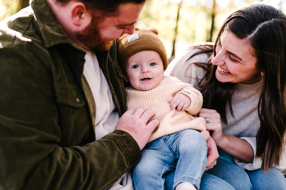 Brunette woman and man sit on either side of their baby son and smile at him