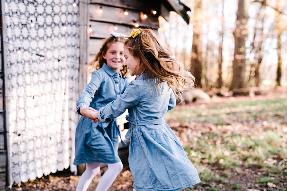 Two young girls in matching denim dresses and hair bows hold hands and swing around in the wood at Lamppost Farm