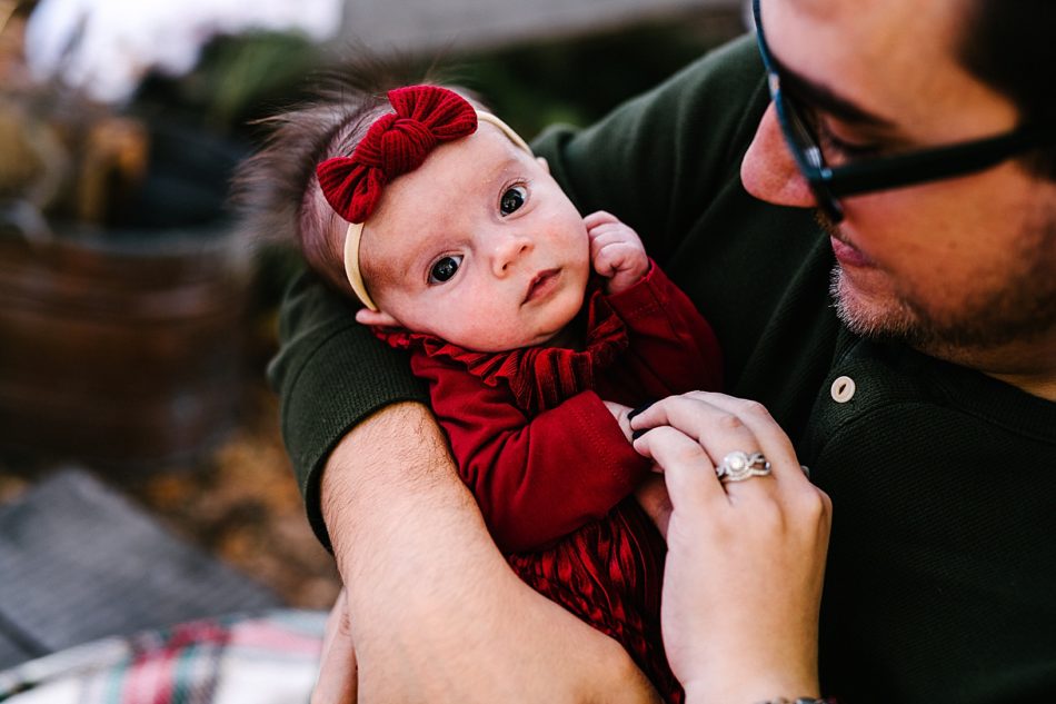 Man holds his newborn baby girl who is wearing a red velvet dress and large red hair bow.