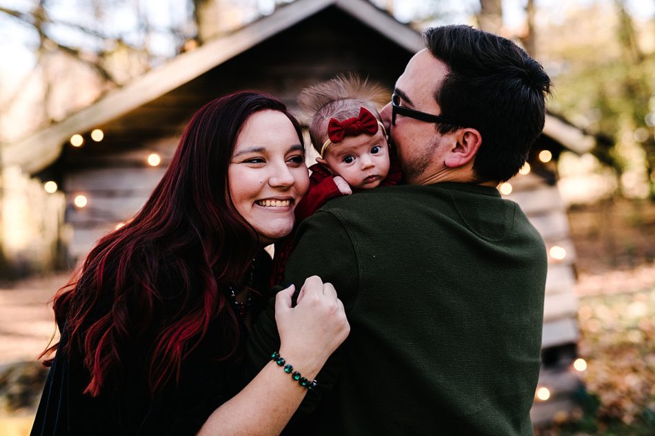 Woman smiles and hugs against husband as his backed is turned to the camera with their newborn peaking over his shoulder.