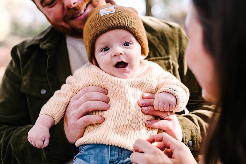 Young baby wearing a mustard colored beanie, an ivory knit sweater, and blue jeans smiles with his mouth open at the camera while being held up by a bearded man.