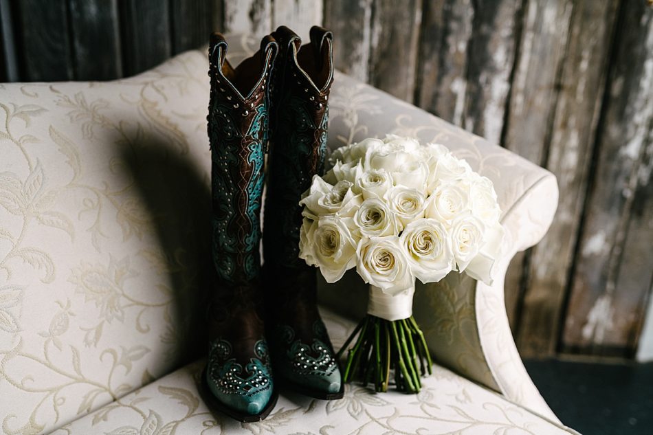 The bride's white rose bouquet wrapped in white silk and her brown and teal studded cowboy boots sitting on a white couch.