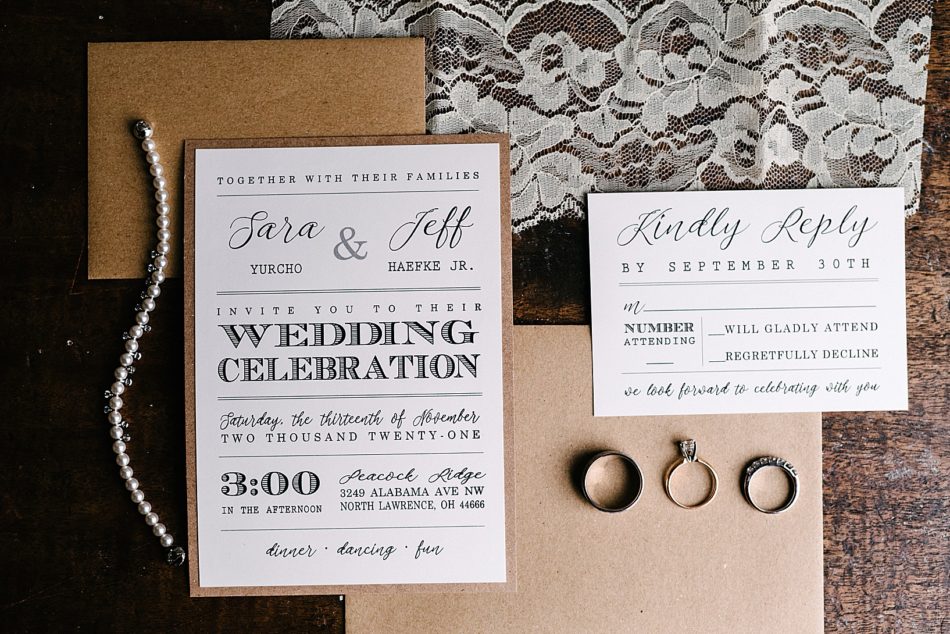 Brown paper and ivory wedding invitations, a string of pearls, a piece of lace, and the bride and grooms wedding rings.
