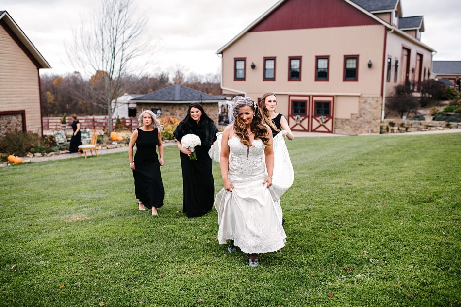 The bride walks through the grass holding the front of her gown while her bridesmaid holds the back of her dress.