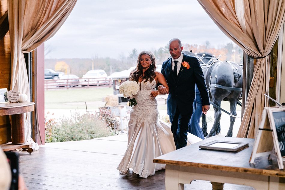 Bride walks arm in arm with her father through giant barn door with curtains drawn on each side.
