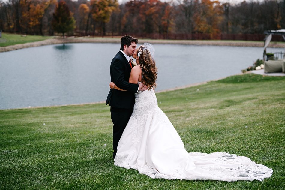 Bride and groom share a kiss on the lawn in front of a small lake at Peacock Ridge with gorgeous fall colored trees lining the background