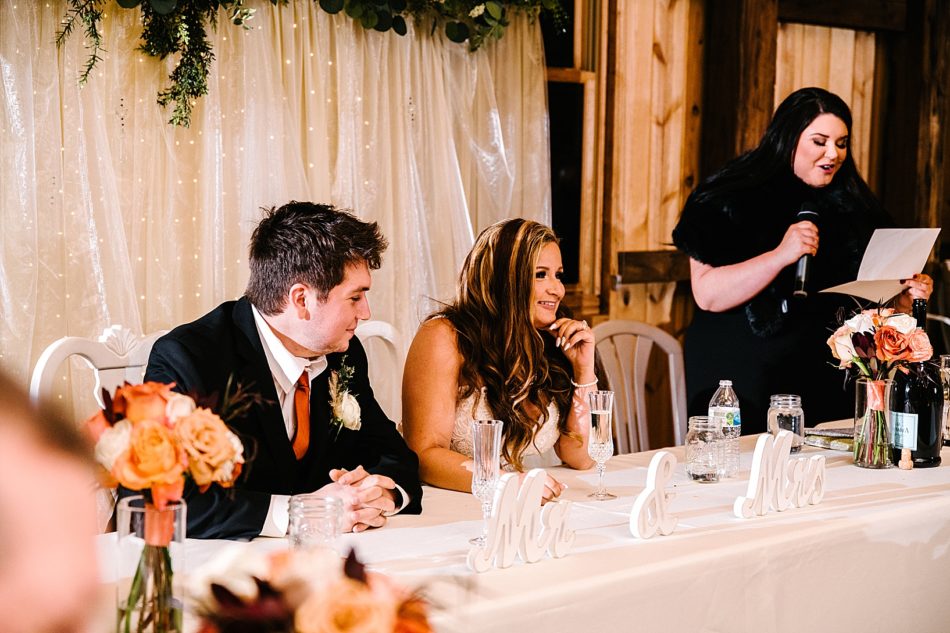 Bride and groom sit at dinner table and smile while maid of honor stands in the background giving speech.