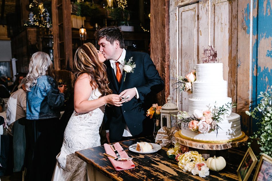 Bride and Groom kiss in front of gorgeous white wedding cake with peach florals and a wooden cake base.