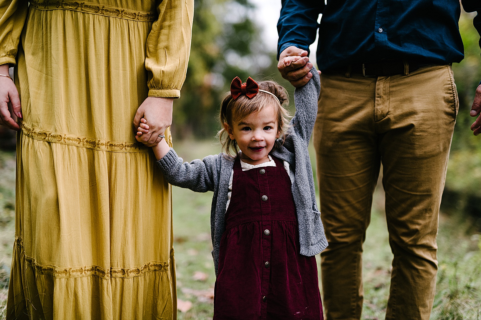 Young girl in a velvet button up dress, matching velvet bow, and grey cardigan holds her parents hands on either side of her.